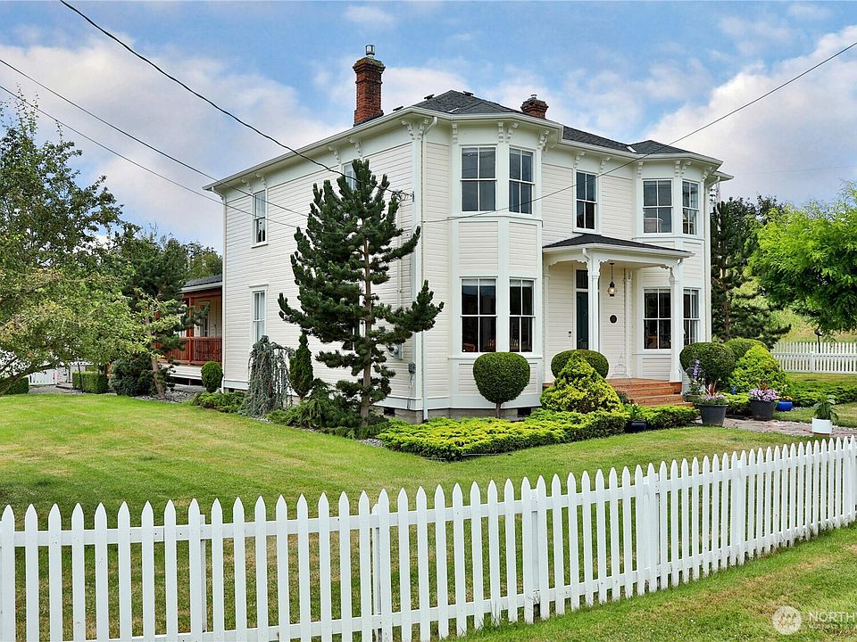 The Victorian residence from the side, framed by mature evergreens with the white picket fence in the foreground
